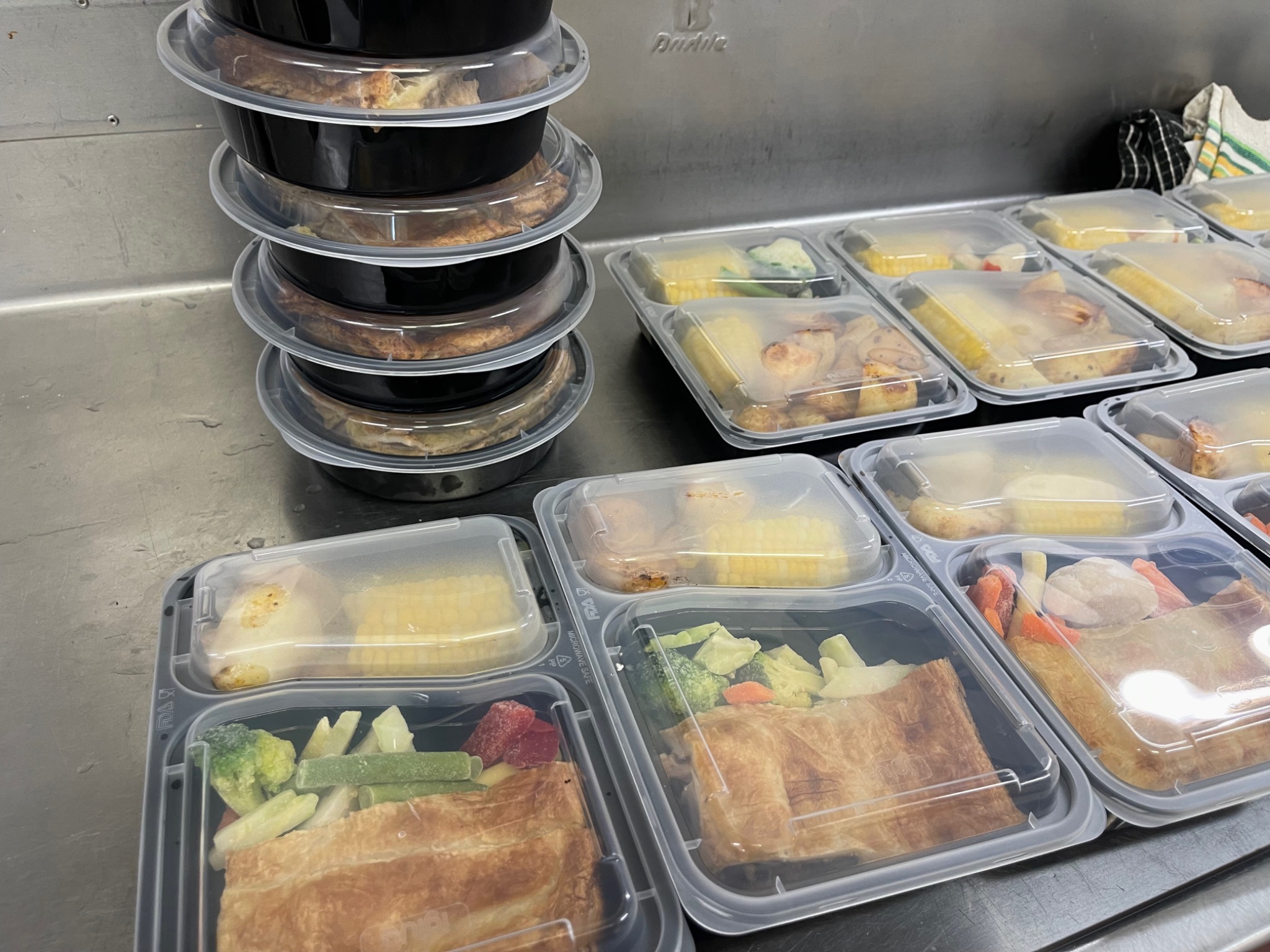 Actual prepared meals in compartmented containers, arranged on a stainless steel bench