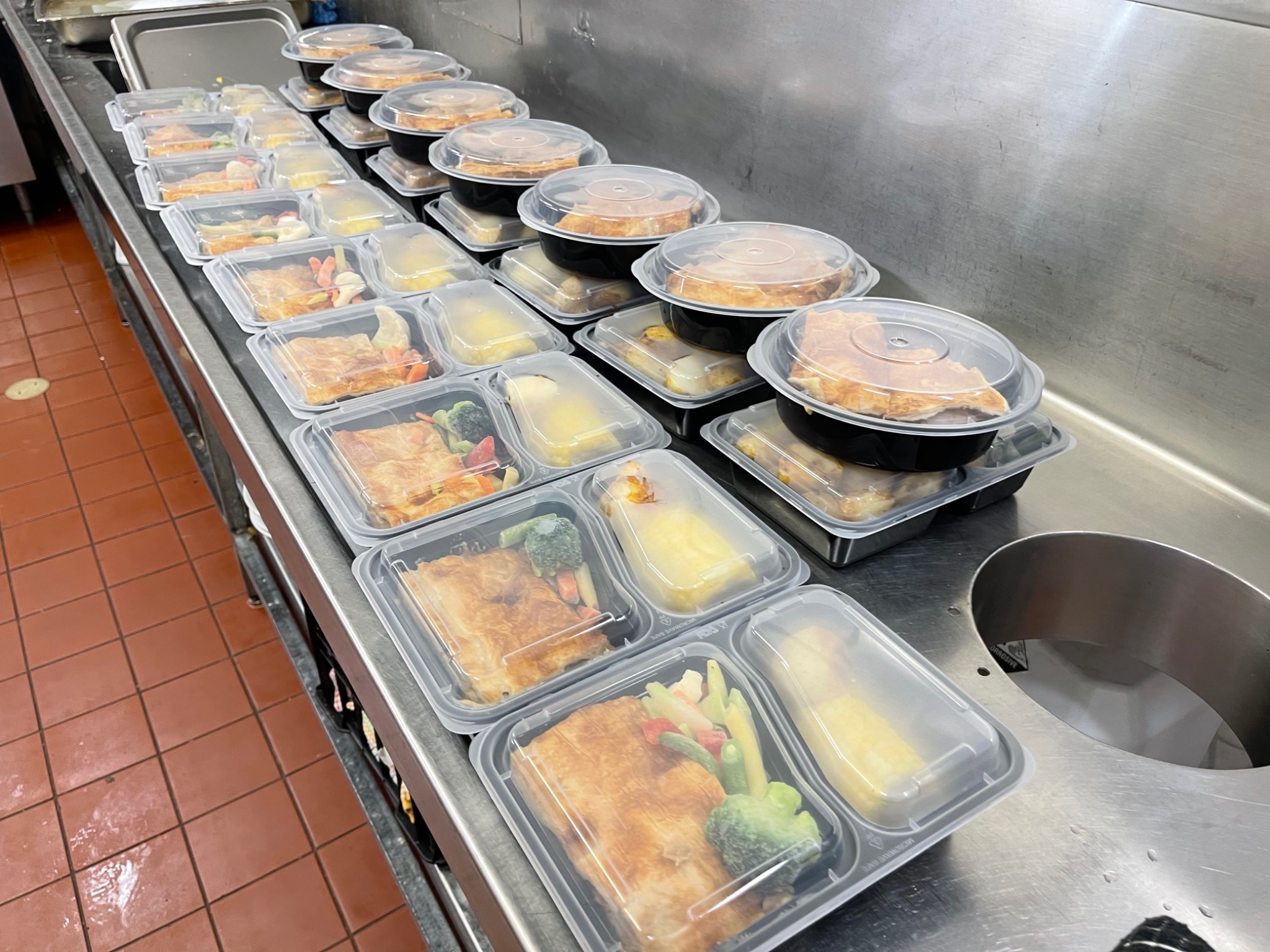 A batch of individual meals packed in trays along a stainless steel kitchen bench